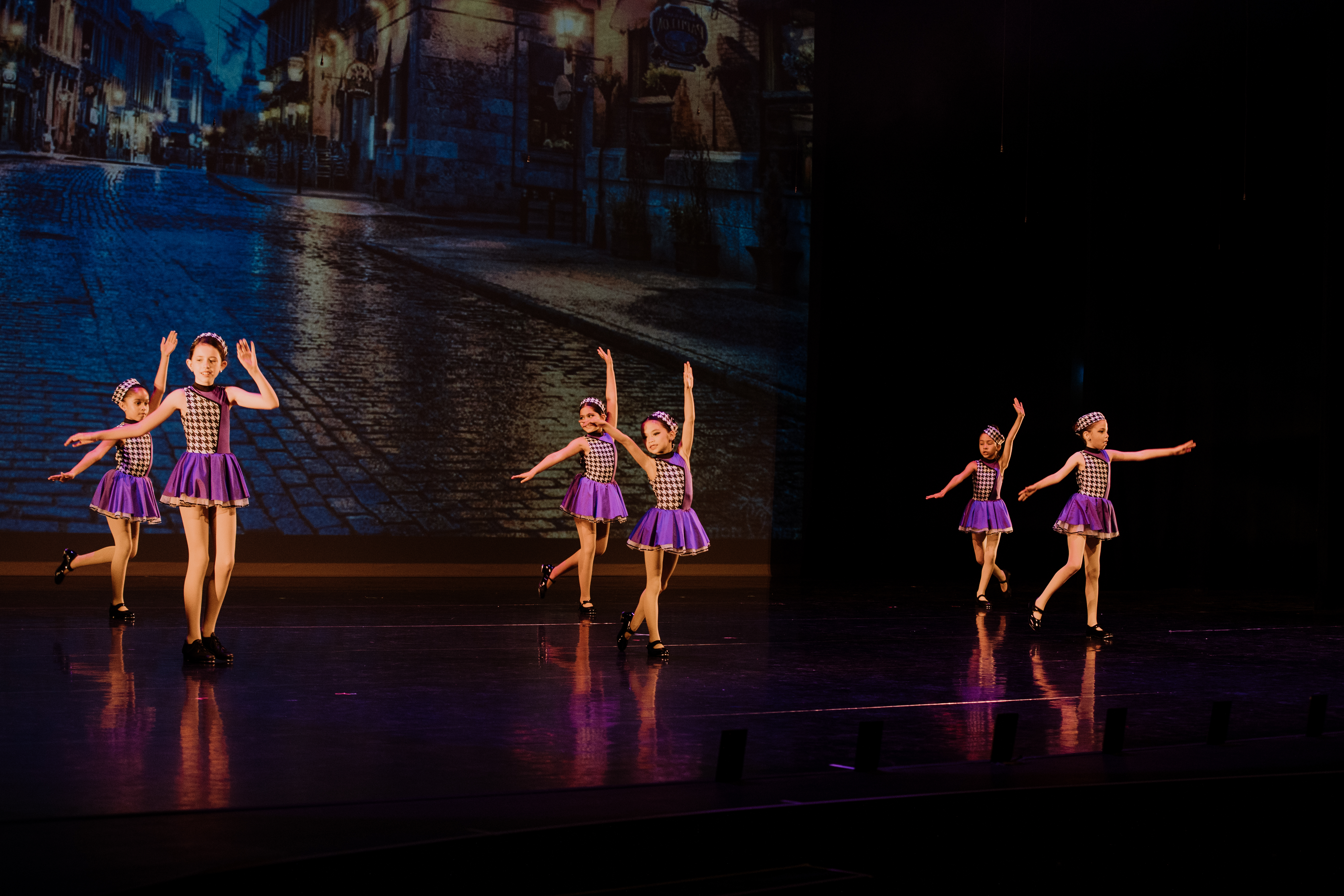 Six dancers performing a tap routine in purple and black and white checkered outfits